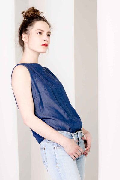 Model lehnt lässig an der Wand in ärmelloser, blauer Bluse. Leicht glänzender Stoff. Nachhaltige Tencel Faser